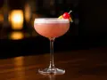 En vackert skummande rosa drink, en Pink Lady, med en körsbärs- och citrondekoration, står på en träbar.