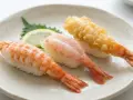 Tre olika sorters sushi med räkor på en tallrik. En med kokt räka, en med rå räka och en med friterad räka. En liten citronskiva och en grön bladgrönsak ligger bredvid. Detta är ett exempel på vad som kan menas med sushi.