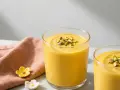 Två glas med krämig indisk yoghurtdryck, toppad med hackade pistagenötter. En rosa servett och små blomformade dekorationer ligger bredvid.