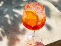 Ett glas med aperol spritz med sprite, is och en apelsinskiva. Skuggor från löv faller över drinken.