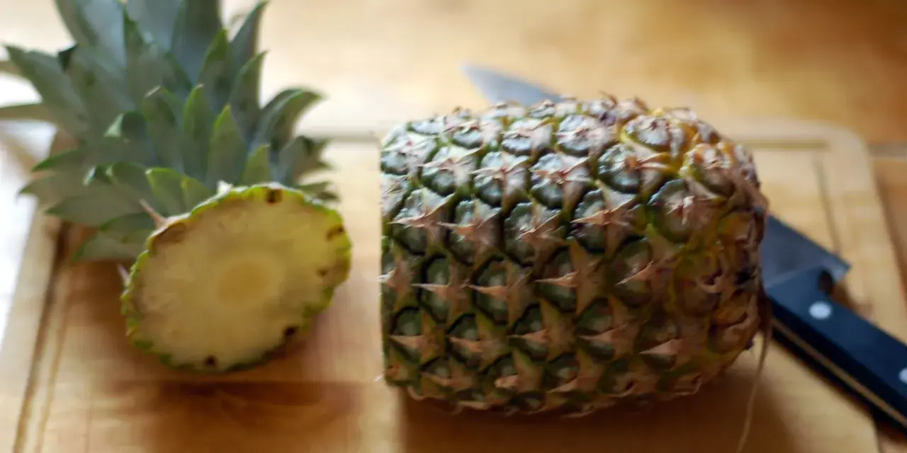 En ananas är delad på mitten på en skärbräda, redo för att visa hur skalar man en ananas. En kniv ligger bredvid.