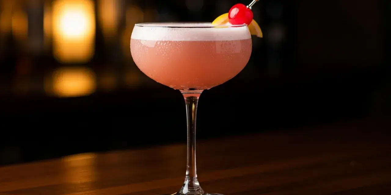 En vackert skummande rosa drink, en Pink Lady, med en körsbärs- och citrondekoration, står på en träbar.