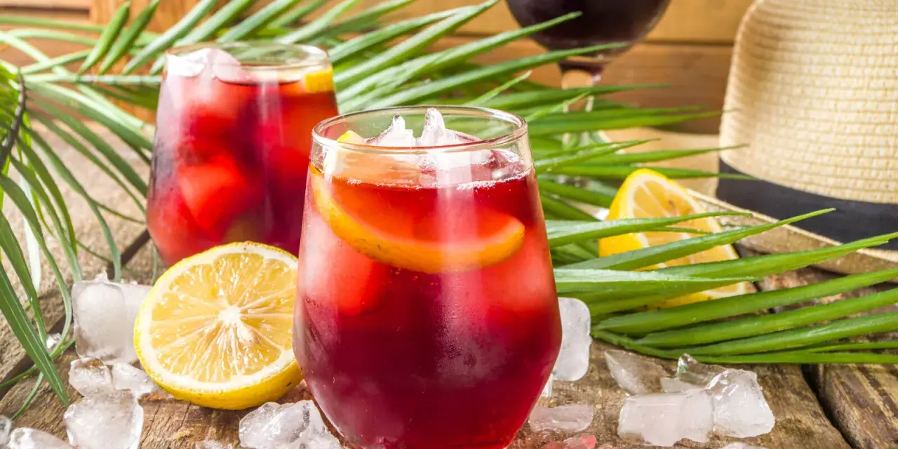 Två glas med tinto de verano, is och citronskivor, omgivna av palmblad och en sommarhatt. Perfekt för en varm dag.