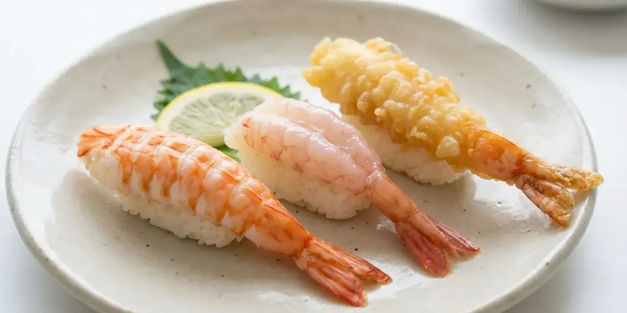 Tre olika sorters sushi med räkor på en tallrik. En med kokt räka, en med rå räka och en med friterad räka. En liten citronskiva och en grön bladgrönsak ligger bredvid. Detta är ett exempel på vad som kan menas med sushi.