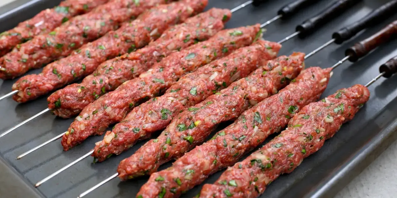 Råa adana kebab-spett med lök och persilja på en plåt, redo för grillen.