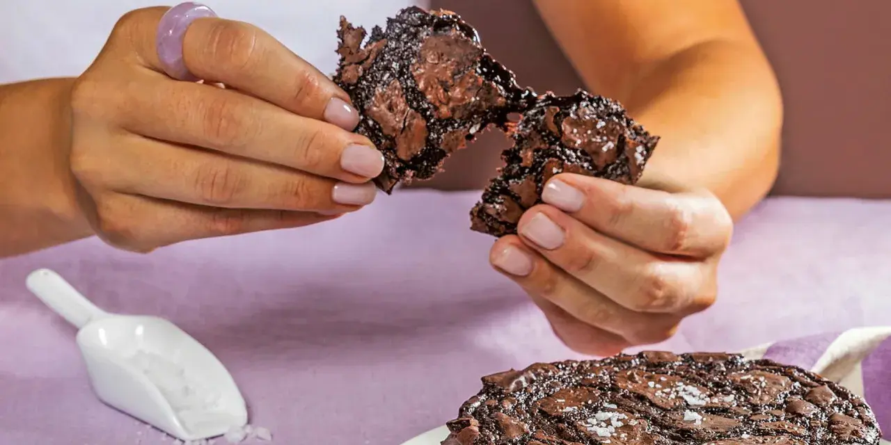 Händerna bryter isär en saftig chokladkaka, strödd med flingsalt. En hel kaka ligger på en tallrik.