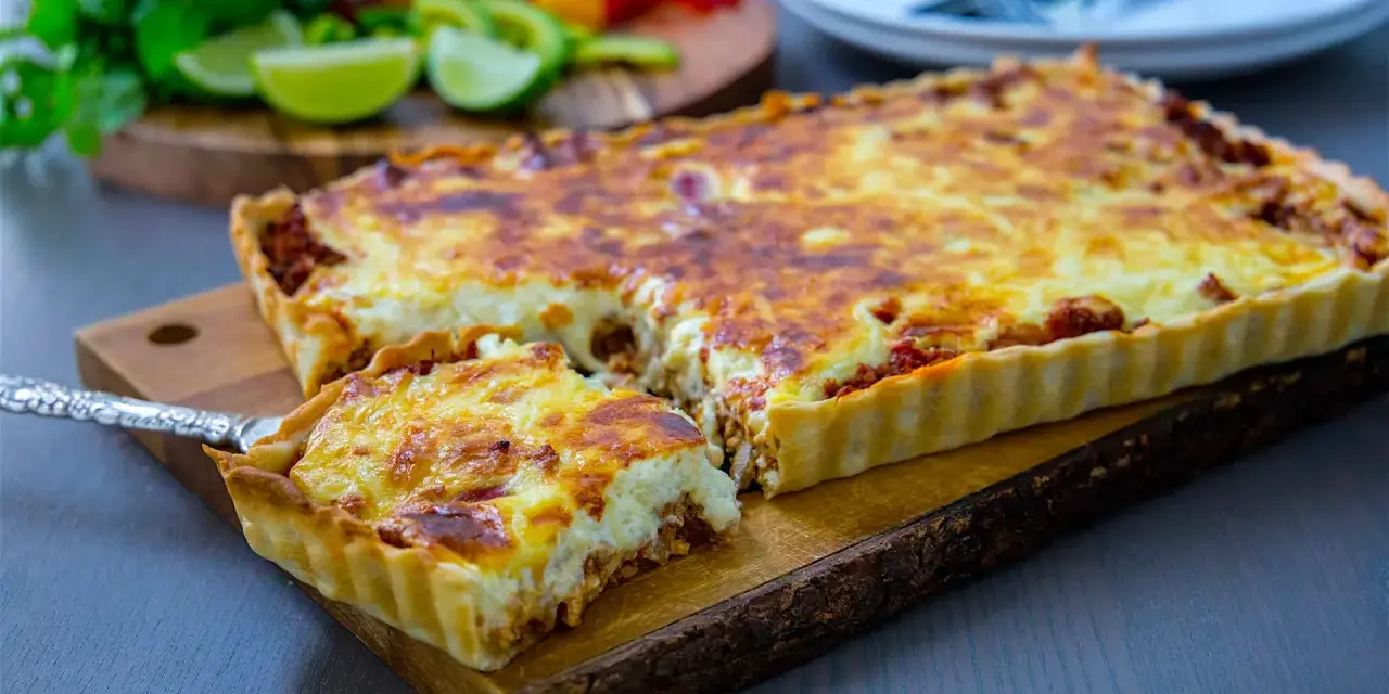En gyllene tacopaj med en krämig fyllning och köttfärs, redo att serveras. Tillbehören med lime och koriander antyder ett smakrikt tacopaj recept.