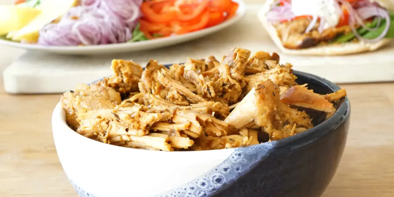 En skål med saftigt gyroskött, redo för ett fantastiskt gyros recept. I bakgrunden syns en sallad och en färdig gyros.