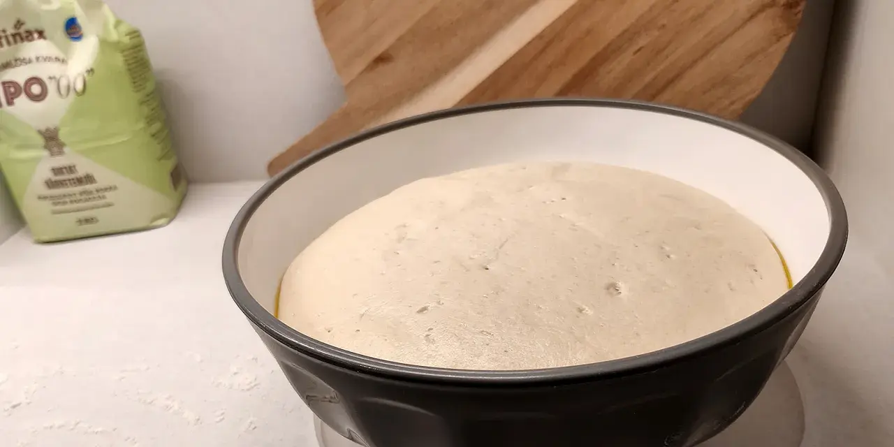 En fluffig pizzadeg i en skål, redo att frysa in för framtida pizzakvällar. En mjölpåse syns i bakgrunden.