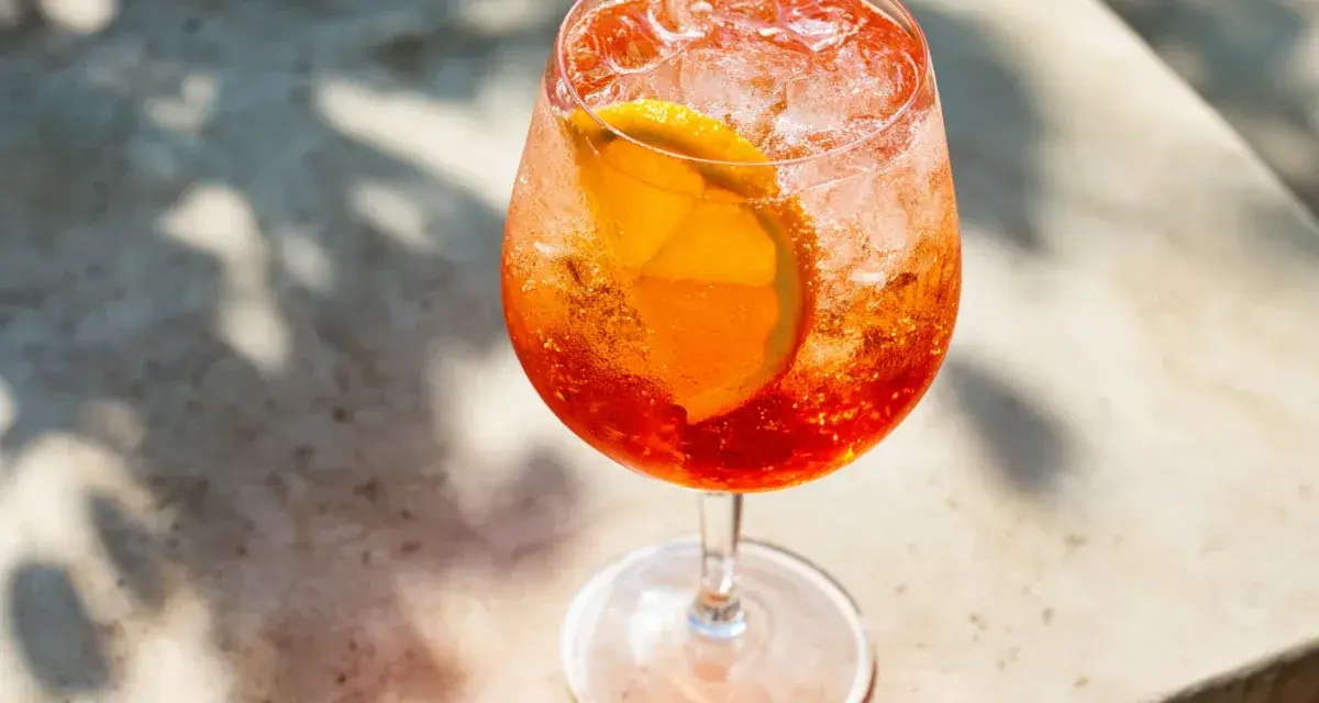 Ett glas med aperol spritz med sprite, is och en apelsinskiva. Skuggor från löv faller över drinken.