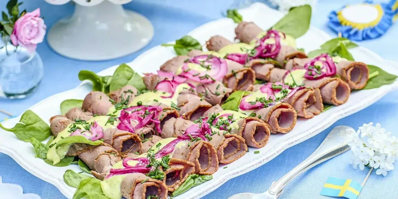Rullad rostbiff med grön sås och rödlök, en perfekt lyxig mat att bjuda på. Serveras på en vit bricka med gröna blad.