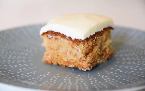 En bit saftig glutenfri morotskaka med krämig glasyr på en gråblå tallrik med prickigt mönster.