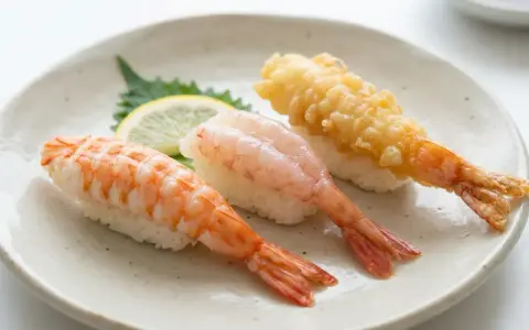 Tre olika sorters sushi med räkor på en tallrik. En med kokt räka, en med rå räka och en med friterad räka. En liten citronskiva och en grön bladgrönsak ligger bredvid. Detta är ett exempel på vad som kan menas med sushi.