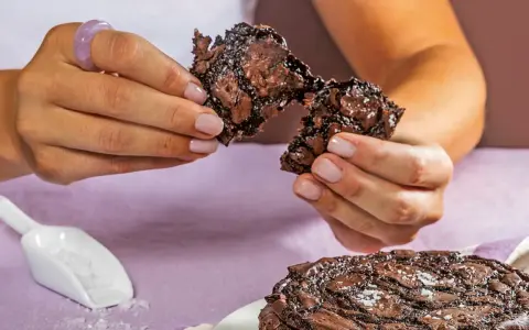 Händerna bryter isär en saftig chokladkaka, strödd med flingsalt. En hel kaka ligger på en tallrik.
