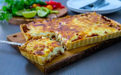 En gyllene tacopaj med en krämig fyllning och köttfärs, redo att serveras. Tillbehören med lime och koriander antyder ett smakrikt tacopaj recept.