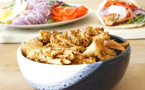 En skål med saftigt gyroskött, redo för ett fantastiskt gyros recept. I bakgrunden syns en sallad och en färdig gyros.