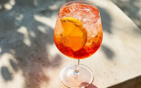 Ett glas med aperol spritz med sprite, is och en apelsinskiva. Skuggor från löv faller över drinken.