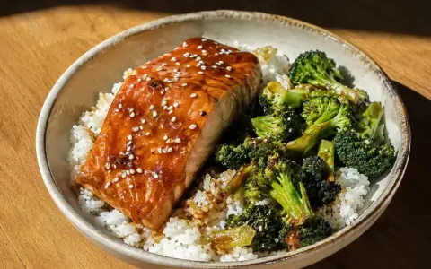 En skål med ris, saftig lax teriyaki toppad med sesamfrön och ångad broccoli.
