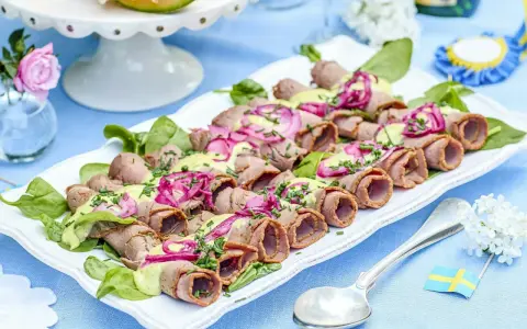 Rullad rostbiff med grön sås och rödlök, en perfekt lyxig mat att bjuda på. Serveras på en vit bricka med gröna blad.