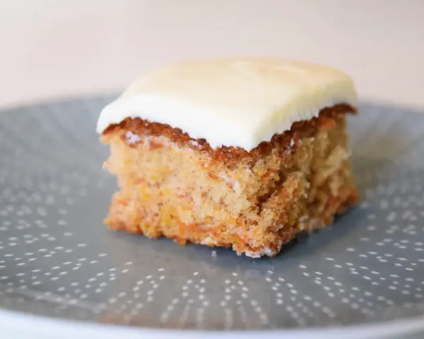 En bit saftig glutenfri morotskaka med krämig glasyr på en gråblå tallrik med prickigt mönster.