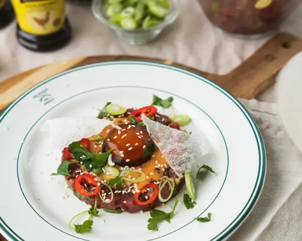 En läcker asiatisk råbiff med chiliringar, salladslök och en krämig sås. Serveras på en vit tallrik med gröna kanter.