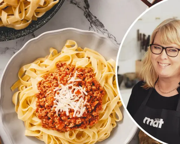 En kvinna med glasögon ler bredvid en skål med tagliatelle och köttfärssås. Perfekt för en snabb middag, den kokta pastan i kylen är redo att serveras.