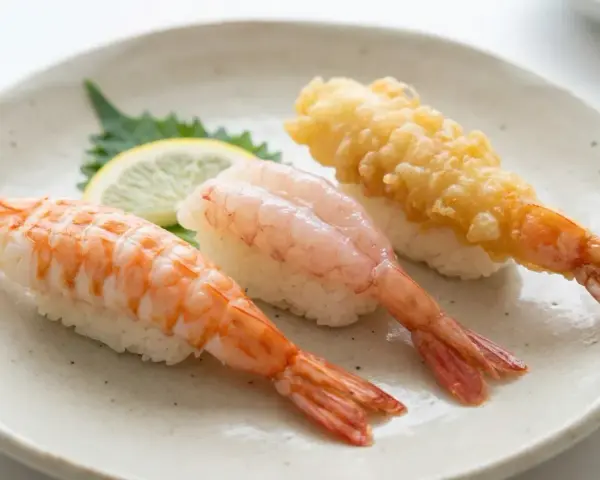 Tre olika sorters sushi med räkor på en tallrik. En med kokt räka, en med rå räka och en med friterad räka. En liten citronskiva och en grön bladgrönsak ligger bredvid. Detta är ett exempel på vad som kan menas med sushi.
