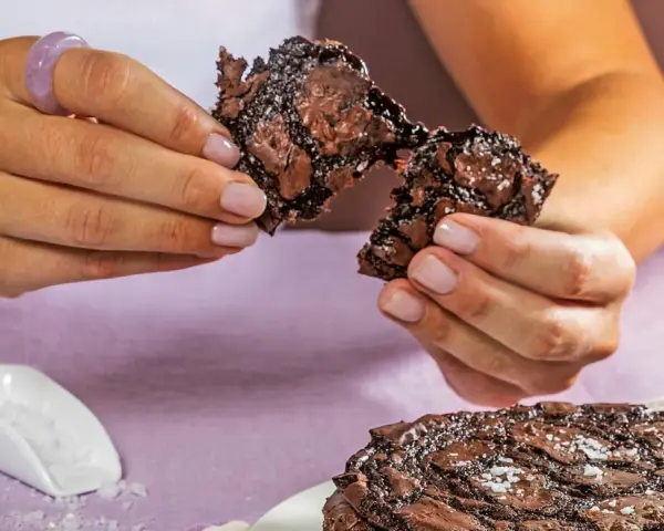 Händerna bryter isär en saftig chokladkaka, strödd med flingsalt. En hel kaka ligger på en tallrik.
