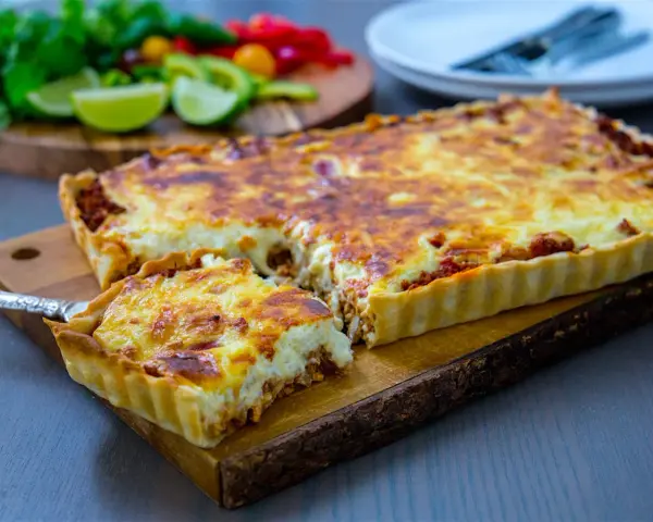 En gyllene tacopaj med en krämig fyllning och köttfärs, redo att serveras. Tillbehören med lime och koriander antyder ett smakrikt tacopaj recept.