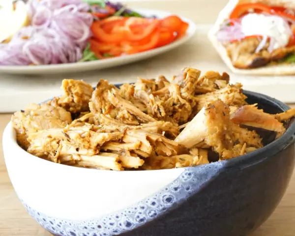 En skål med saftigt gyroskött, redo för ett fantastiskt gyros recept. I bakgrunden syns en sallad och en färdig gyros.