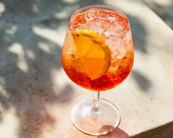 Ett glas med aperol spritz med sprite, is och en apelsinskiva. Skuggor från löv faller över drinken.