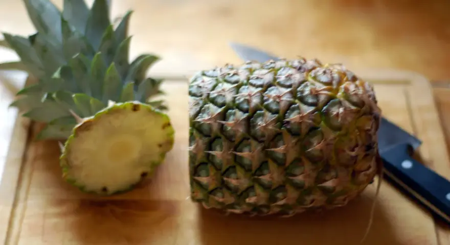 En ananas är delad på mitten på en skärbräda, redo för att visa hur skalar man en ananas. En kniv ligger bredvid.