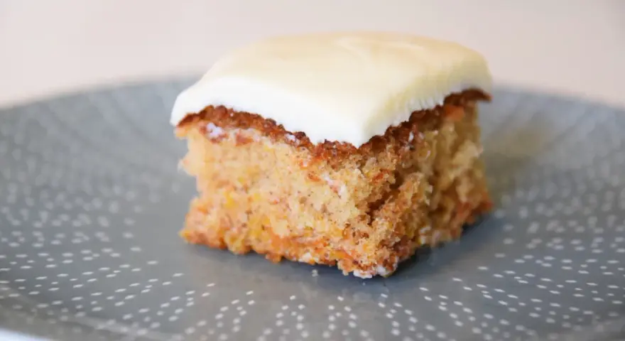 En bit saftig glutenfri morotskaka med krämig glasyr på en gråblå tallrik med prickigt mönster.