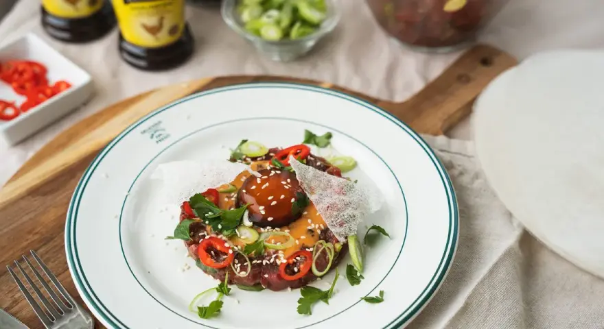 En läcker asiatisk råbiff med chiliringar, salladslök och en krämig sås. Serveras på en vit tallrik med gröna kanter.