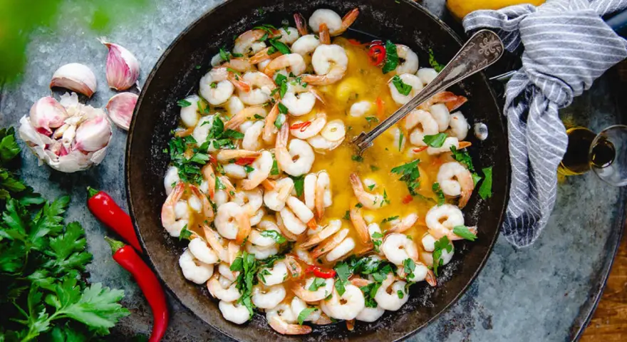 En panna med saftiga räkor i en gyllene sås, perfekt för ett snabbt gambas recept. Garnerad med färsk persilja, chili och vitlök.
