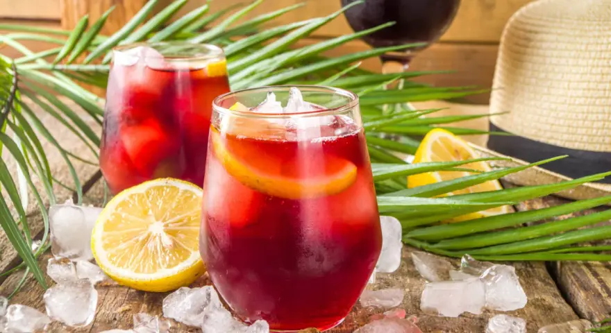 Två glas med tinto de verano, is och citronskivor, omgivna av palmblad och en sommarhatt. Perfekt för en varm dag.