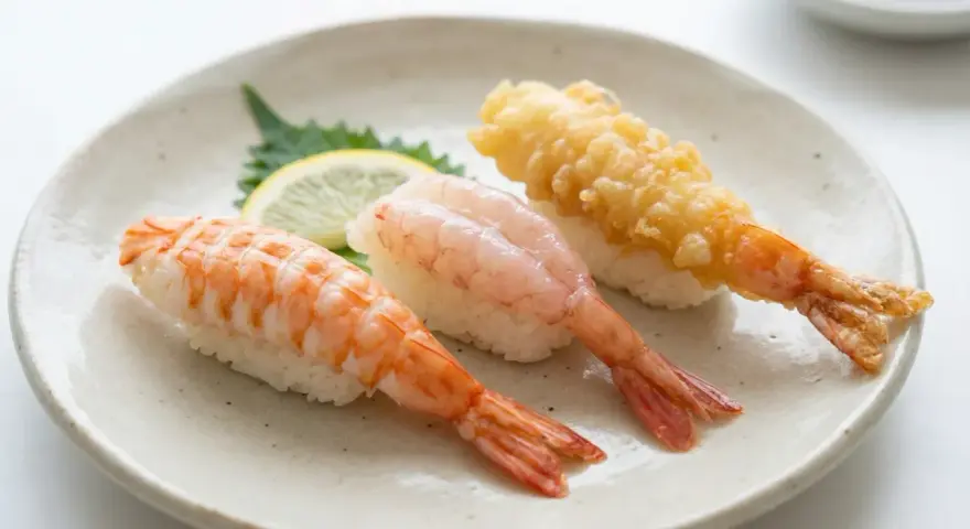 Tre olika sorters sushi med räkor på en tallrik. En med kokt räka, en med rå räka och en med friterad räka. En liten citronskiva och en grön bladgrönsak ligger bredvid. Detta är ett exempel på vad som kan menas med sushi.