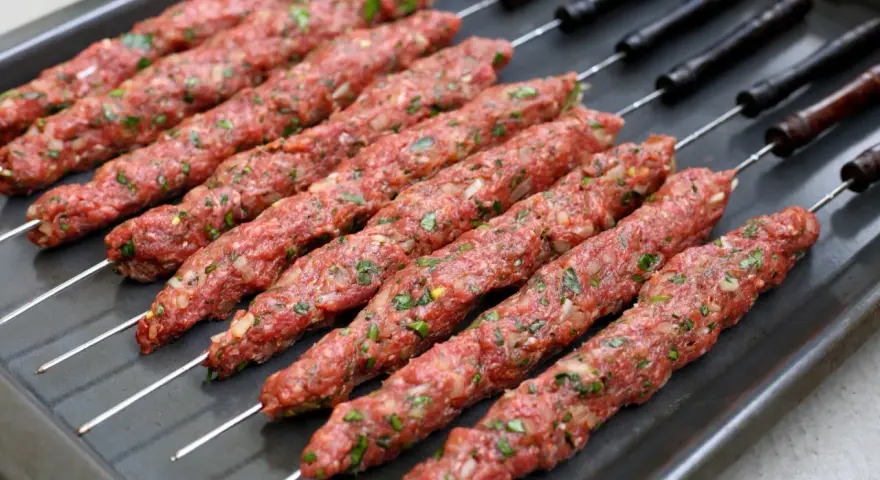 Råa adana kebab-spett med lök och persilja på en plåt, redo för grillen.