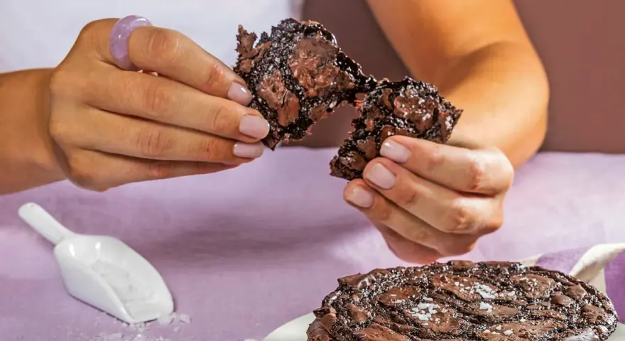 Händerna bryter isär en saftig chokladkaka, strödd med flingsalt. En hel kaka ligger på en tallrik.
