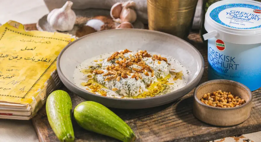 En tallrik med grekisk yoghurt i varm mat, toppad med rostade pinjenötter och kryddor. Bredvid syns squash, vitlök och en gammal kokbok.