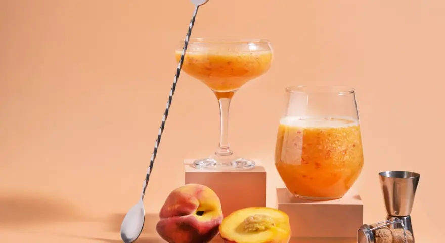Två glas med persikojuoma, persikor och en barsked. Ett enkelt bellini recept för en solig dag.