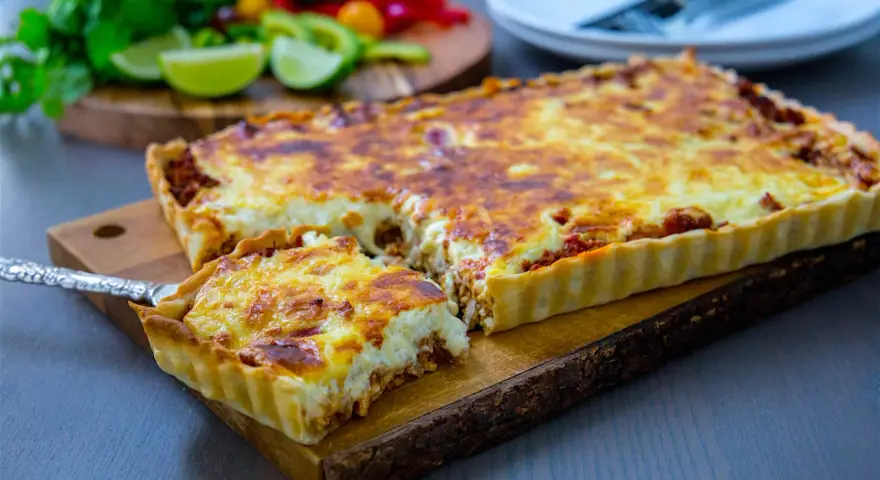 En gyllene tacopaj med en krämig fyllning och köttfärs, redo att serveras. Tillbehören med lime och koriander antyder ett smakrikt tacopaj recept.