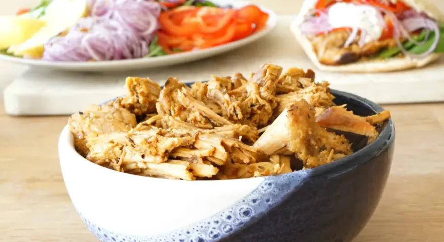 En skål med saftigt gyroskött, redo för ett fantastiskt gyros recept. I bakgrunden syns en sallad och en färdig gyros.