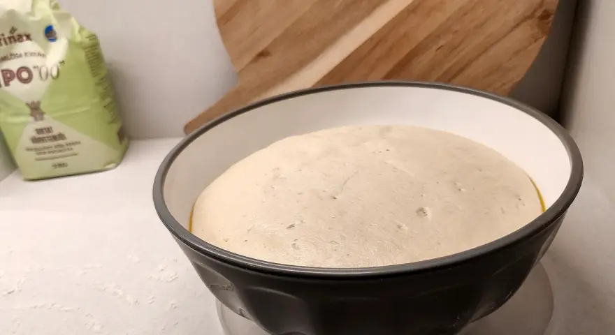 En fluffig pizzadeg i en skål, redo att frysa in för framtida pizzakvällar. En mjölpåse syns i bakgrunden.