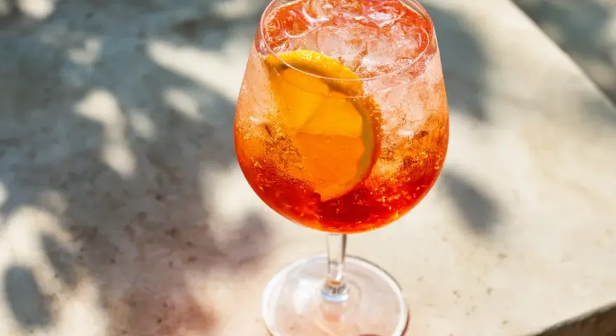 Ett glas med aperol spritz med sprite, is och en apelsinskiva. Skuggor från löv faller över drinken.