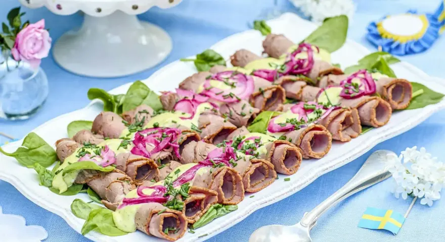 Rullad rostbiff med grön sås och rödlök, en perfekt lyxig mat att bjuda på. Serveras på en vit bricka med gröna blad.