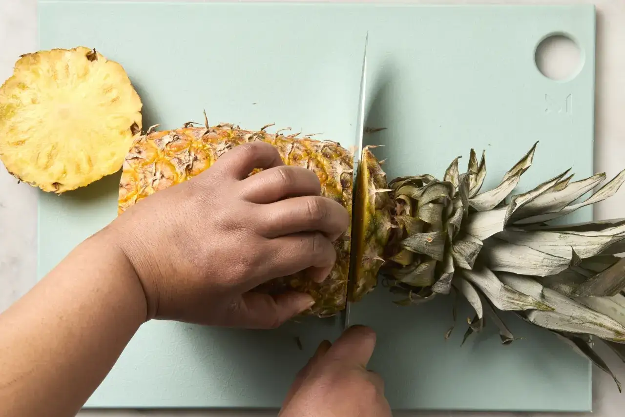 Här visas hur skalar man en ananas. En kniv skär genom den taggiga frukten på en ljusblå skärbräda. En skiva ananas ligger bredvid.