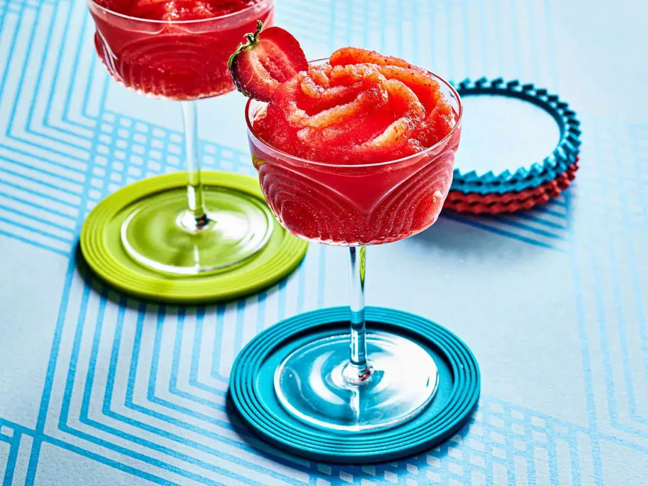 Tv&aring; glas med fryst jordgubbsdaiquiri, en garnerad med en jordgubbe, st&aring;r p&aring; f&auml;rgglada underl&auml;gg.