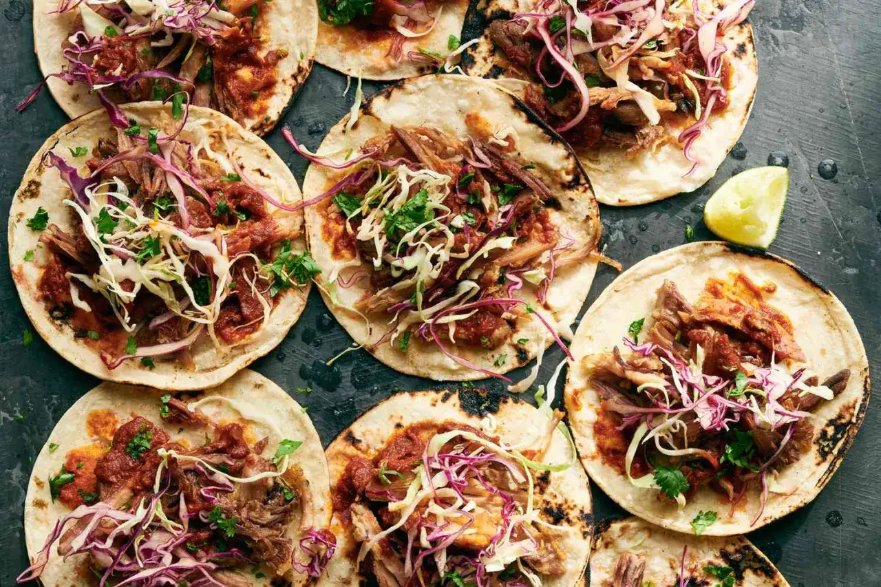 Flera tacos med saftigt carnitas recept, toppade med krispig k&aring;l och f&auml;rsk koriander. En limeklyfta ligger bredvid.