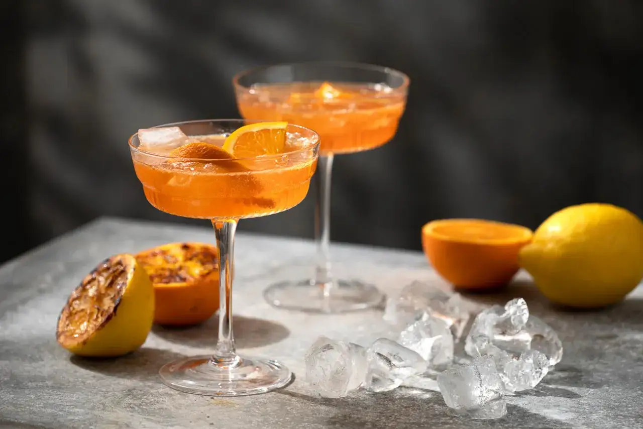 Tv&aring; glas med aperol spritz med sprite, garnerade med apelsinskivor. Isbitar och citrusfrukter ligger utspridda.